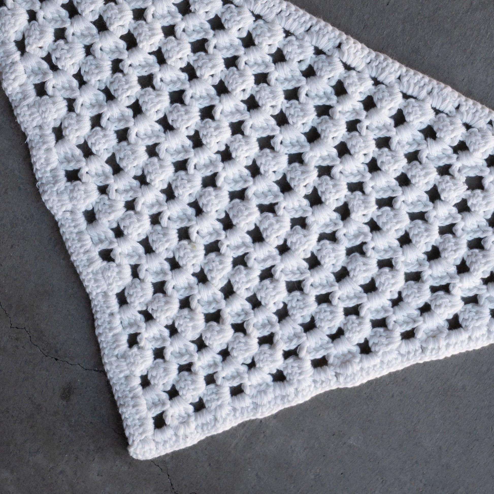The Knit Head Scarf | Cloud by forever french baby features a white crocheted triangle design with a lacy, open pattern, crafted from soft cotton yarn and displayed on a gray textured surface. An adjustable tie ensures a comfortable and customizable fit.