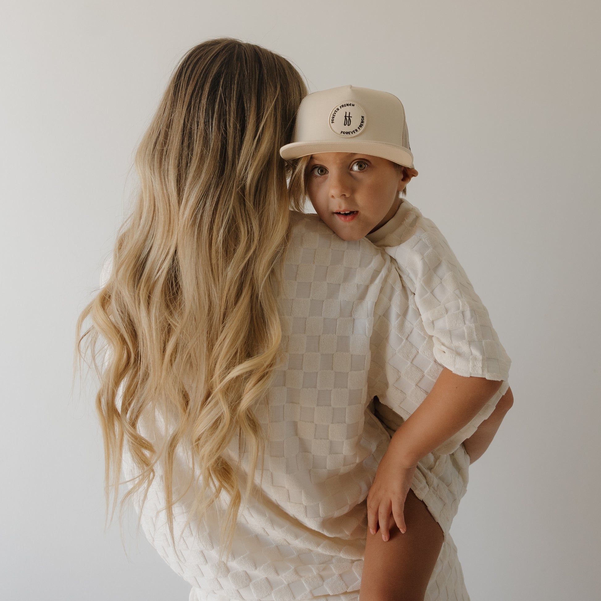 A woman with long, wavy blonde hair holds a young child dressed in the forever french baby Child Terry Short Set in Coconut Milk and a matching cap. The child faces the camera while the woman looks away, both against a light backdrop.