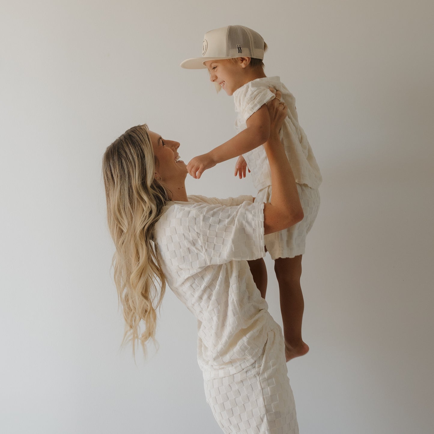A woman with long blonde hair lifts a smiling young child in a beige cap, both wearing coordinated textured cotton shorts from the forever french baby Child Terry Short Set in Coconut Milk, against a plain white background.