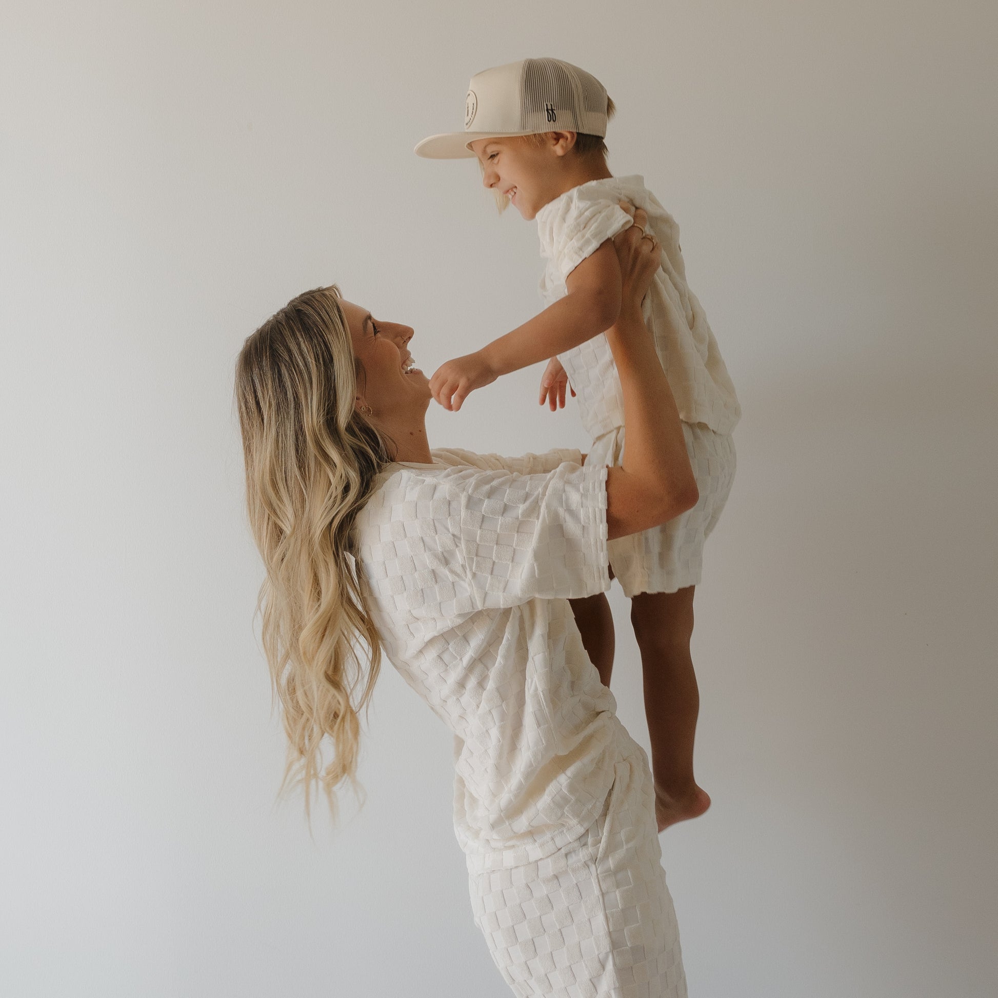 A woman with long blonde hair lifts a smiling young child in a beige cap, both wearing coordinated textured cotton shorts from the forever french baby Child Terry Short Set in Coconut Milk, against a plain white background.