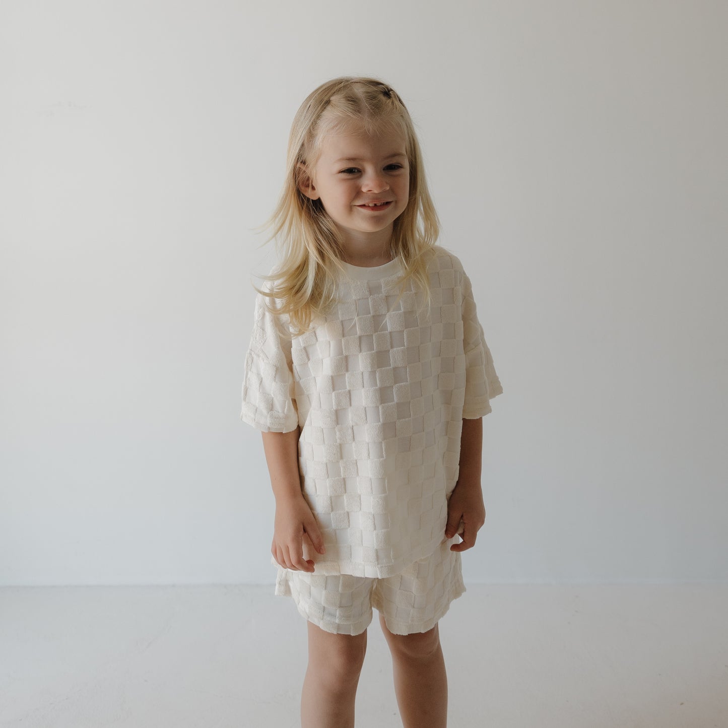 A young child with long blonde hair smiles in the forever french baby Child Terry Short Set in Coconut Milk, featuring matching shorts, against a plain white background.