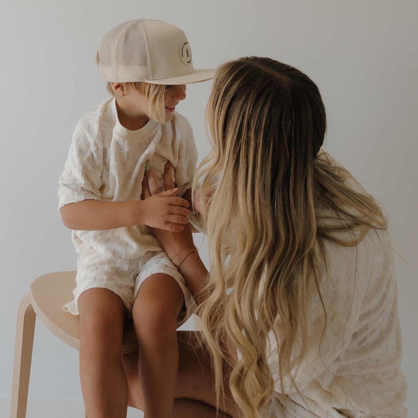 A young child in a forever french baby Child Terry Short Set in Coconut Milk and a beige hat sits on a wooden stool, smiling at a woman with long blonde hair wearing matching family outfits as they interact warmly.