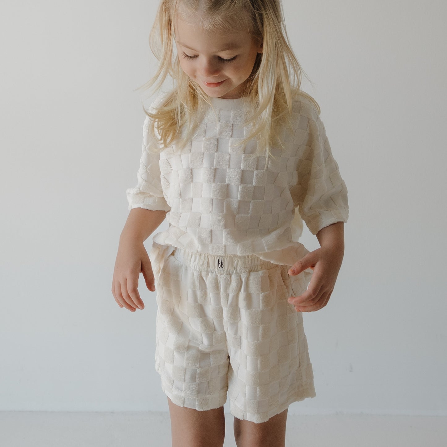 A young child with long blonde hair wears the forever french baby Child Terry Short Set in Coconut Milk, standing against a white background and looking down with a slight smile—ideal for family matching outfits.