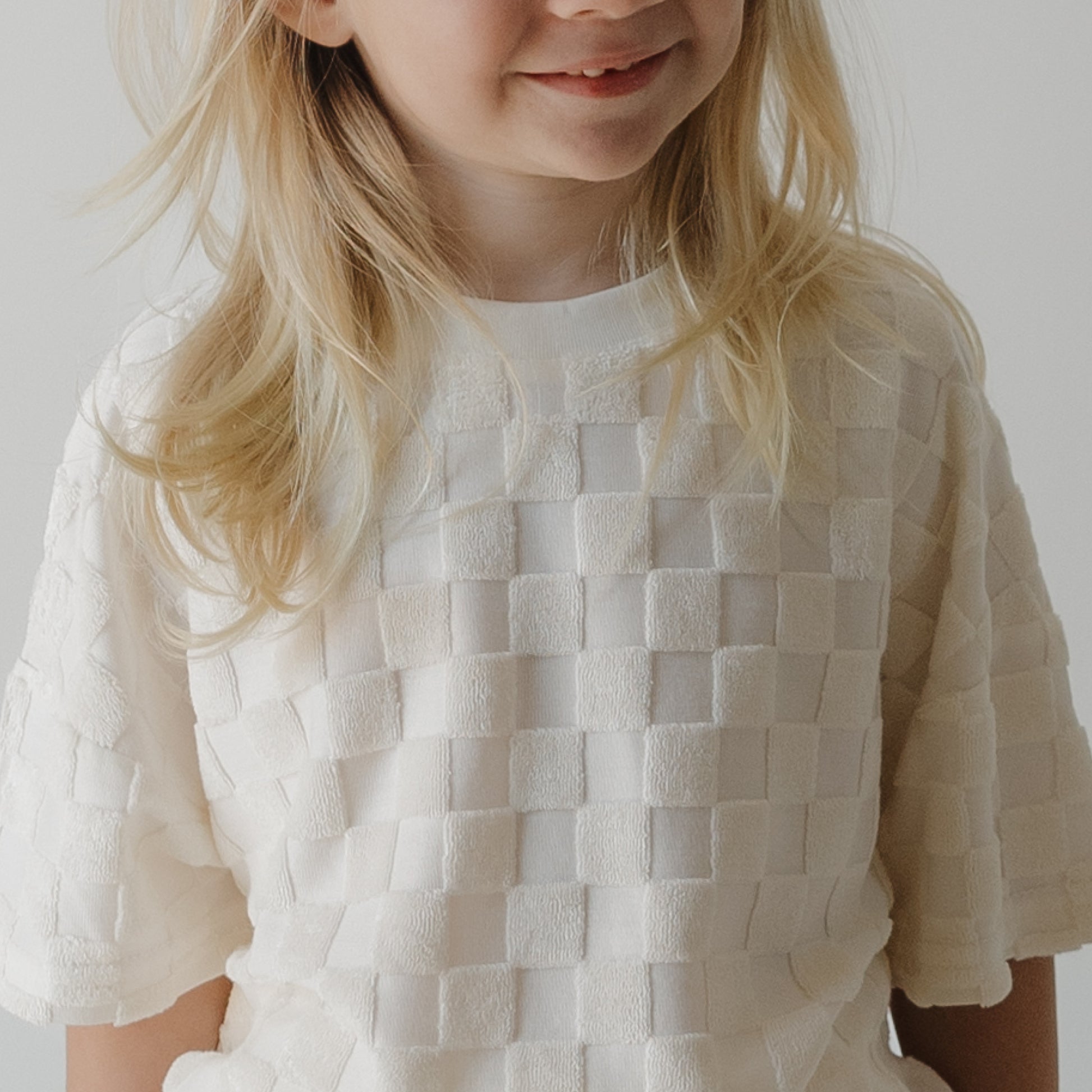 A young child with long blonde hair wears the forever french baby Child Terry Short Set in Coconut Milk, smiling softly against a plain light background.
