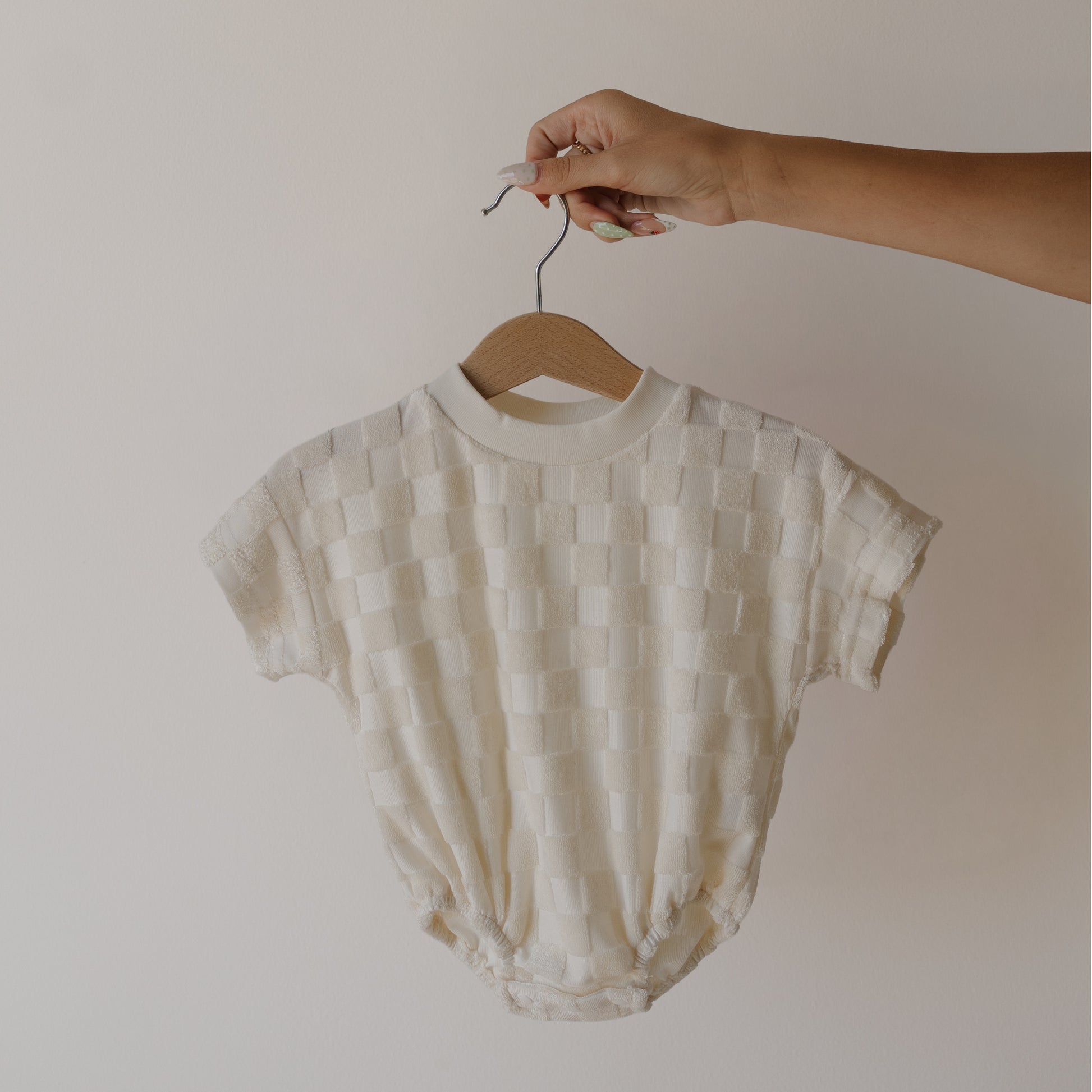 A hand holds the forever french baby Terry T-Shirt Romper in Coconut Milk, a short-sleeved, cream romper with a textured checkerboard pattern—ideal for coordinating family outfits—against a simple light background.