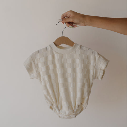 A hand holds the forever french baby Terry T-Shirt Romper in Coconut Milk, a short-sleeved, cream romper with a textured checkerboard pattern—ideal for coordinating family outfits—against a simple light background.