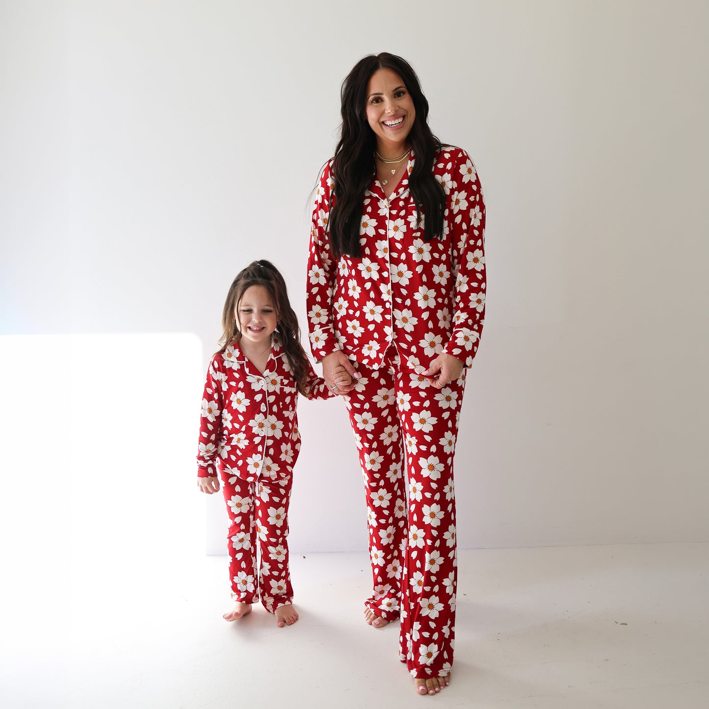 A woman and a young girl stand barefoot, holding hands and smiling, both wearing matching forever french baby Women's Bamboo Button Up Lounge Sets in the Cherry Blossoms print, posing against a plain white background.