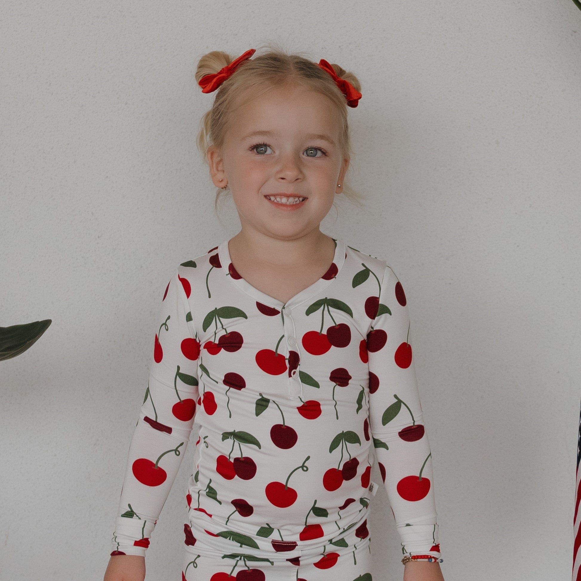 A young girl with blonde hair in two buns with red bows smiles, wearing forever french baby Bamboo Two Piece Pajamas | Cherry, featuring a red cherry print. She stands against a plain light-colored wall.