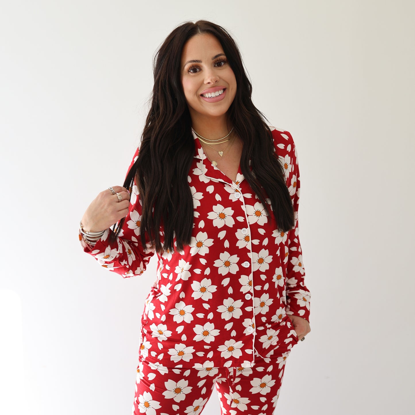 A woman with long dark hair smiles wearing the forever french baby Women's Bamboo Button Up Lounge Set | Cherry Blossoms, featuring red pajamas with a white daisy floral pattern, against a plain white background.