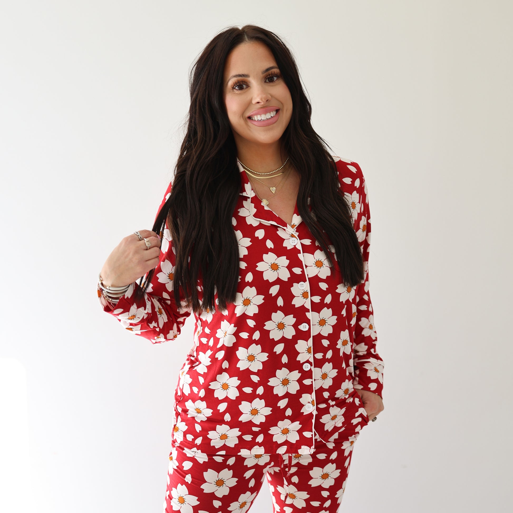 A woman with long dark hair smiles wearing the forever french baby Women's Bamboo Button Up Lounge Set | Cherry Blossoms, featuring red pajamas with a white daisy floral pattern, against a plain white background.