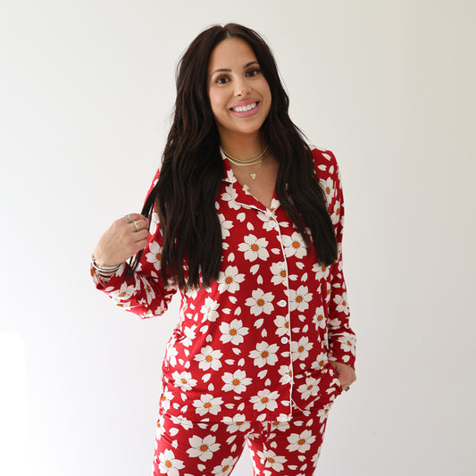 A woman with long dark hair smiles wearing the forever french baby Women's Bamboo Button Up Lounge Set | Cherry Blossoms, featuring red pajamas with a white daisy floral pattern, against a plain white background.