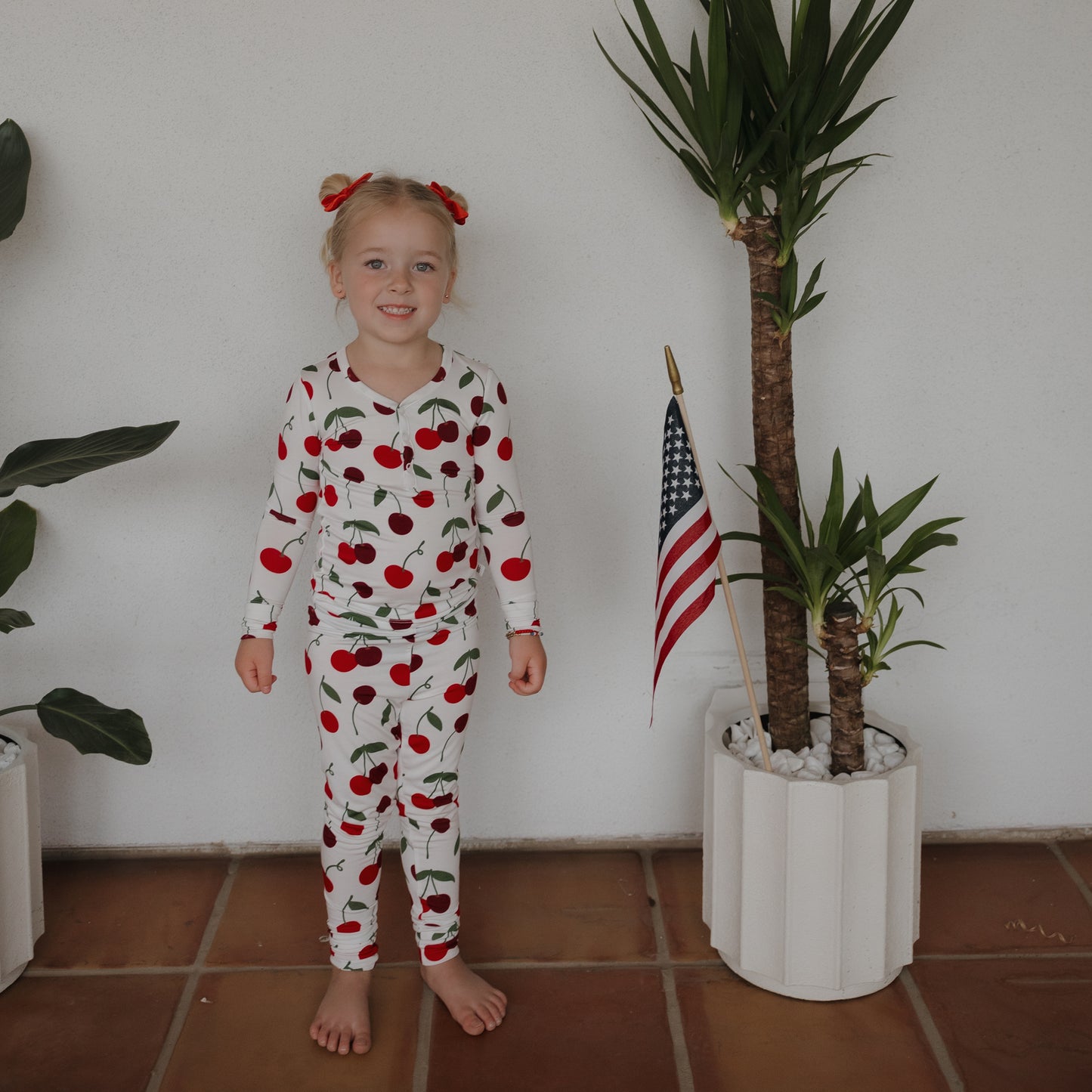A young girl smiles barefoot beside a potted plant with an American flag, wearing forever french baby Bamboo Two Piece Pajamas | Cherry. White walls and lush plants surround her, highlighting the hypo-allergenic kids sleepwear.