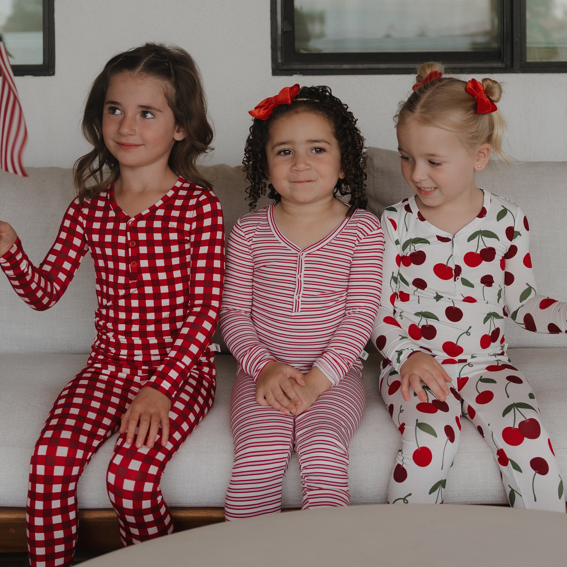 Three young girls wearing forever french baby Bamboo Two Piece Pajamas—one in red and white checks, one in pink stripes, and one in cherry print—sit smiling on a couch with red hair bows.