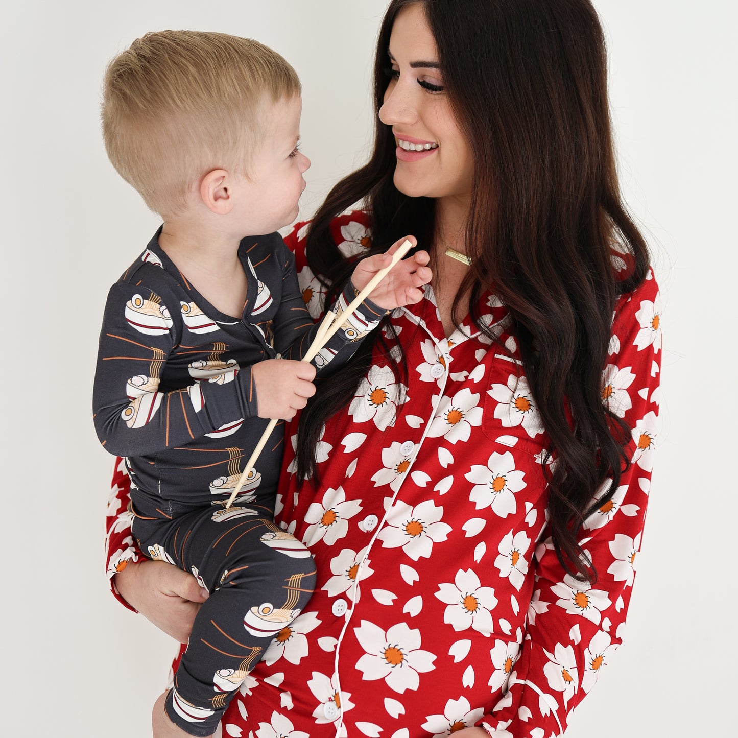 A woman in red floral bamboo pajamas smiles at a child dressed in forever french baby Bamboo Two Piece Pajama | Ramen with a fun cartoon print, both sharing a happy moment against a plain white background.