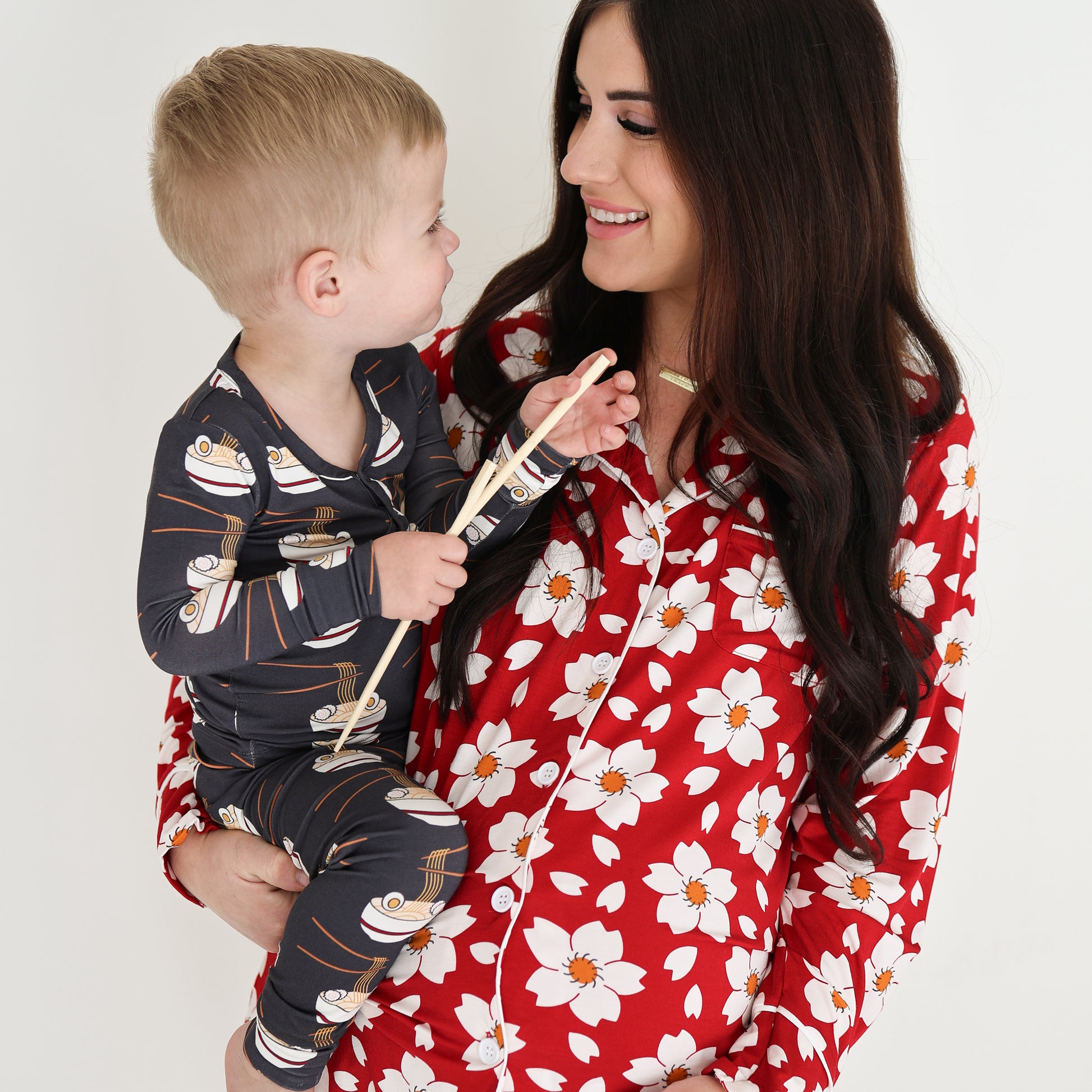 A woman in red floral bamboo pajamas smiles at a child dressed in forever french baby Bamboo Two Piece Pajama | Ramen with a fun cartoon print, both sharing a happy moment against a plain white background.