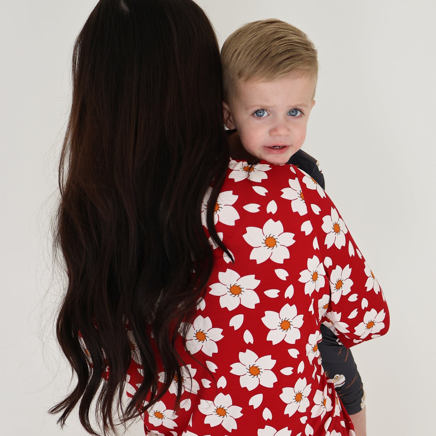 A woman with long dark hair in a red floral dress holds a young blond boy wearing forever french baby Bamboo Two Piece Pajama | Ramen, as he looks over her shoulder toward the camera against a plain white background.