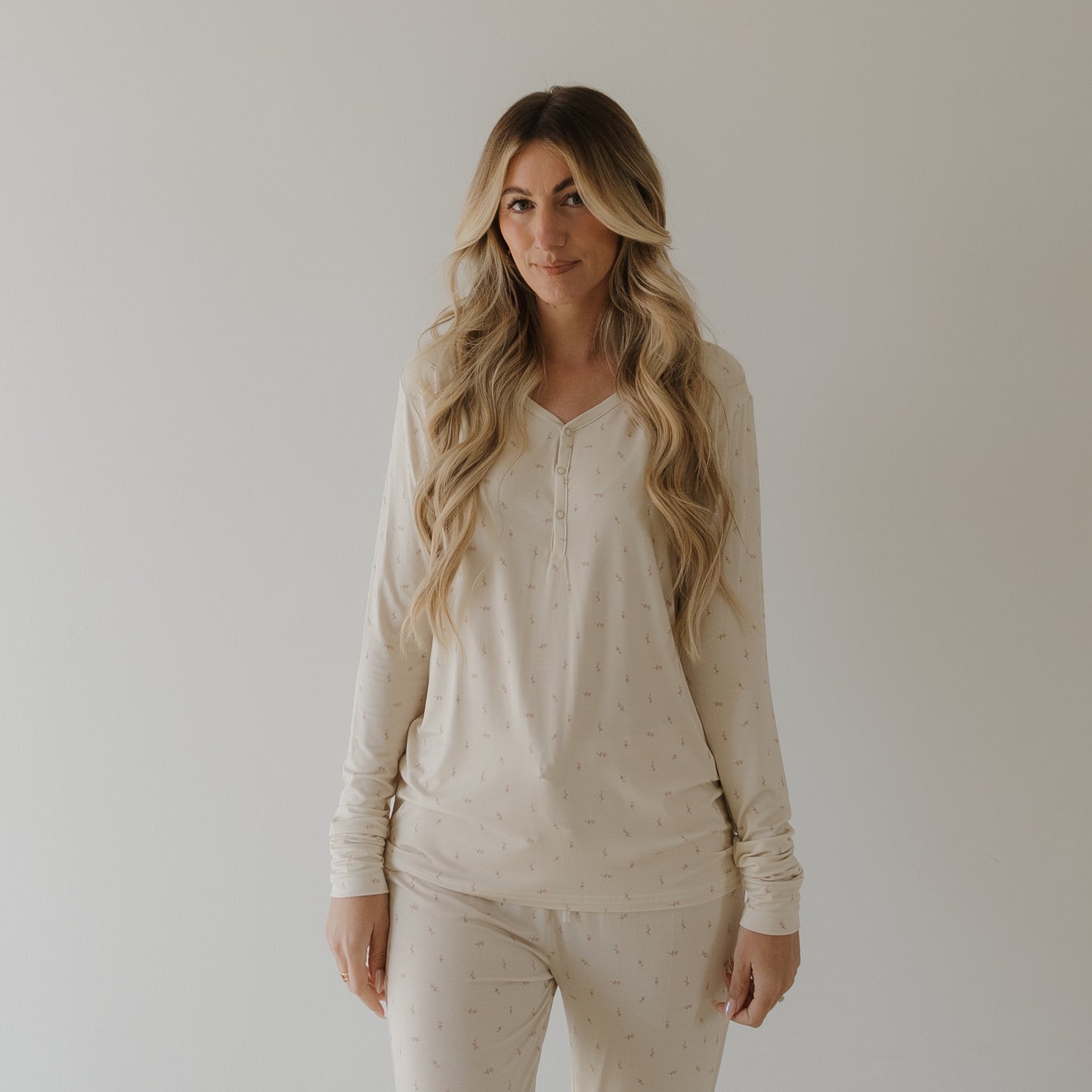 A woman with long, wavy blond hair stands against a white background, wearing the forever french baby Women's Long Sleeve Bamboo Pajama | Lucy set featuring a subtle dotted pattern.