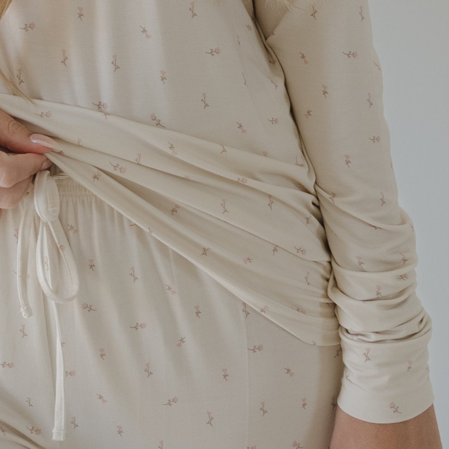 A person wearing the forever french baby Women's Long Sleeve Bamboo Pajama | Lucy in a cream color with pink floral patterns lifts the hem to show its drawstring waistband; only their torso and one hand are visible.