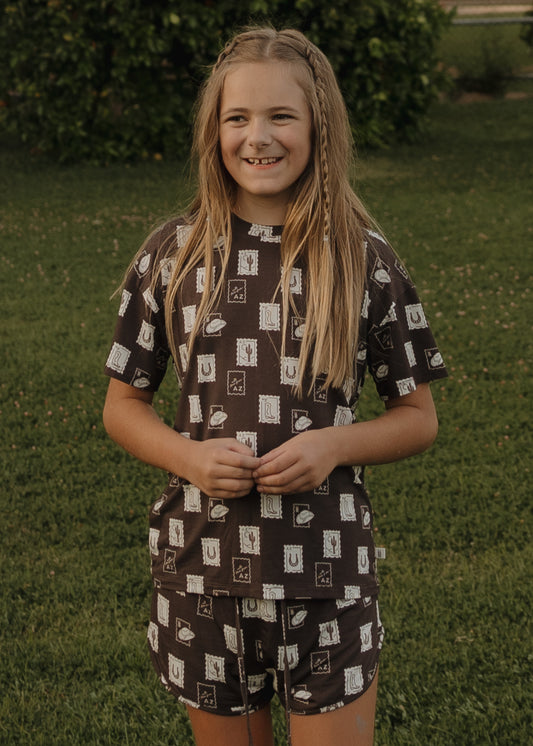 A young girl with long blonde hair smiles on the grass, wearing the forever french baby Girl's Bamboo Short Set from With Love from AZ. The comfy set features breathable bamboo fabric and a light geometric print, with bushes blurred in the background.