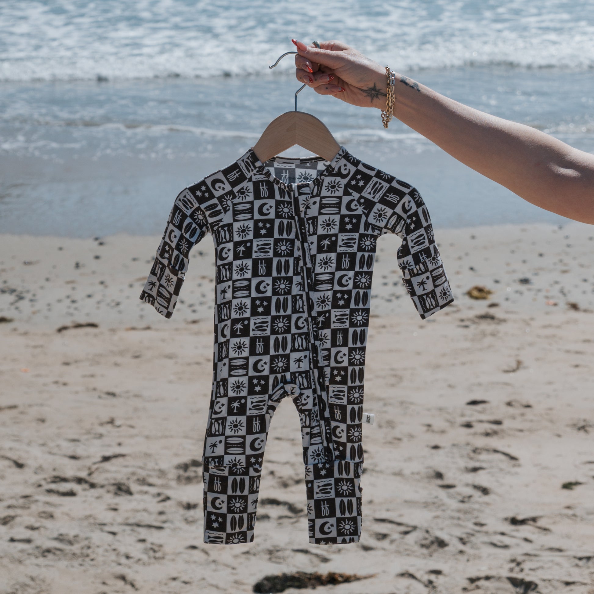 A hand holds a hanger displaying the forever french baby Bamboo Zip Pajamas | Summer Check, a black and white patterned, breathable baby sleepwear jumpsuit, on a sandy beach with the ocean and waves in the background.