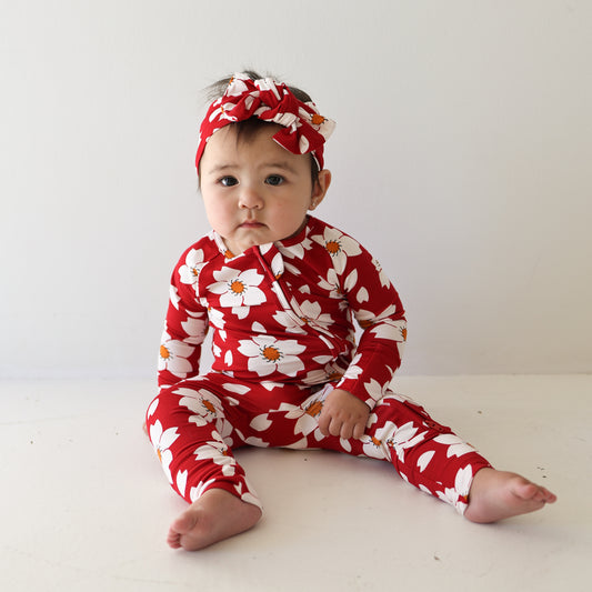 A baby sits on the floor against a plain white background, wearing forever french baby's Bamboo Zip Pajamas in the Cherry Blossoms print with a matching headband, looking slightly pensive.
