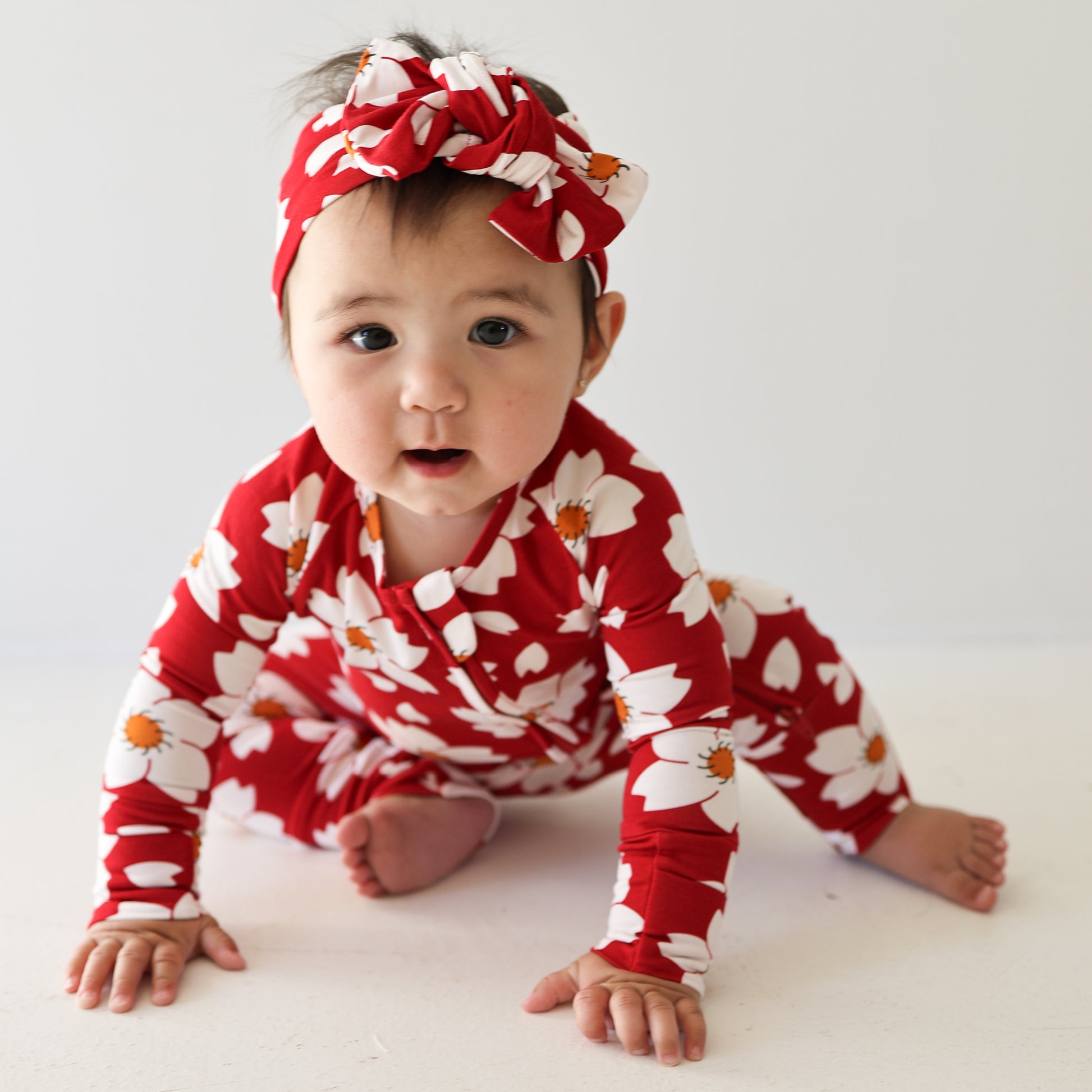 A baby sits on the floor wearing forever french baby's Bamboo Zip Pajamas | Cherry Blossoms—a red outfit with a matching headband featuring a white and orange floral pattern—looking toward the camera against a plain white background.