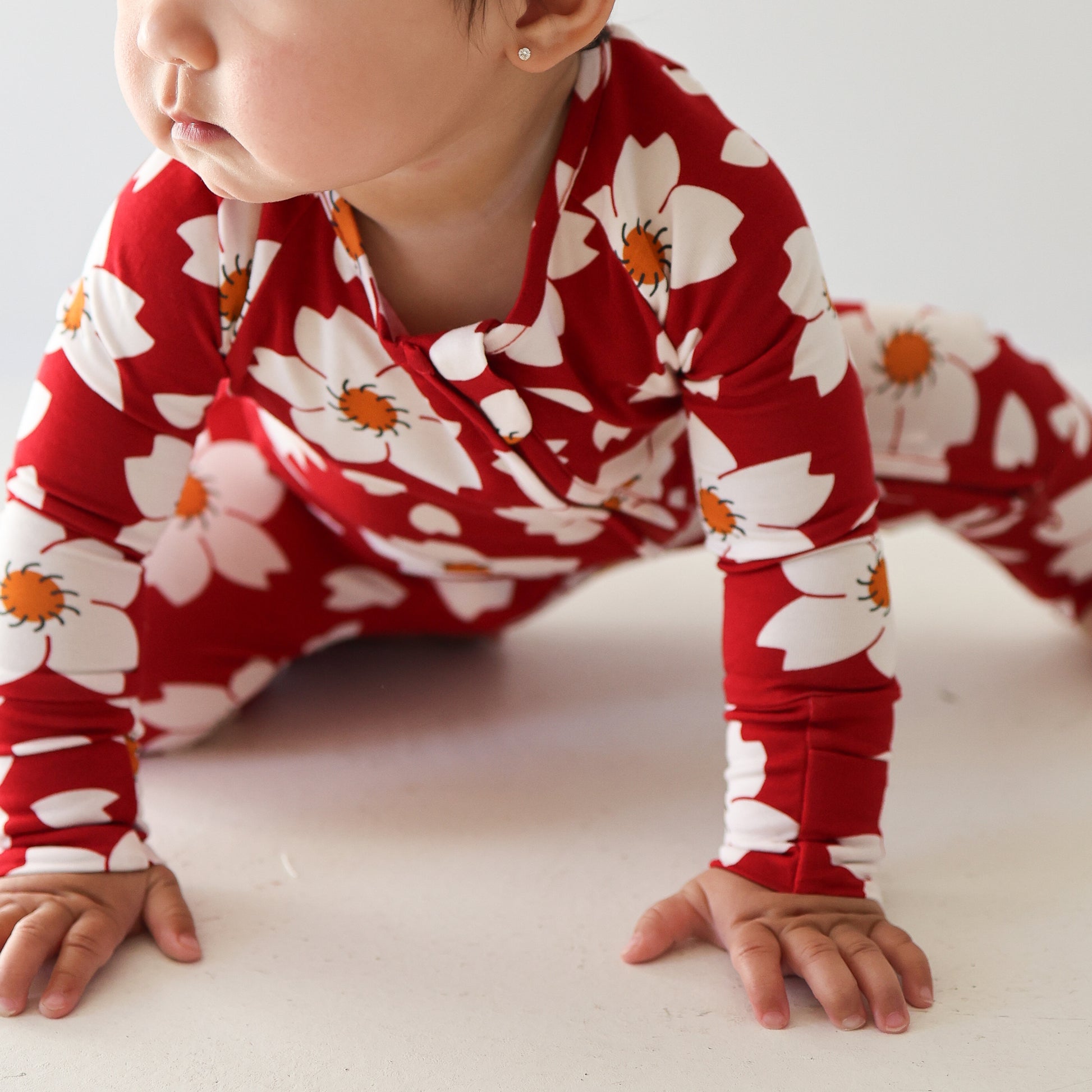 A baby in forever french baby Bamboo Zip Pajamas | Cherry Blossoms, featuring a red outfit with large white and orange flower prints, crawls sideways on a light surface with hands and knees on the floor.