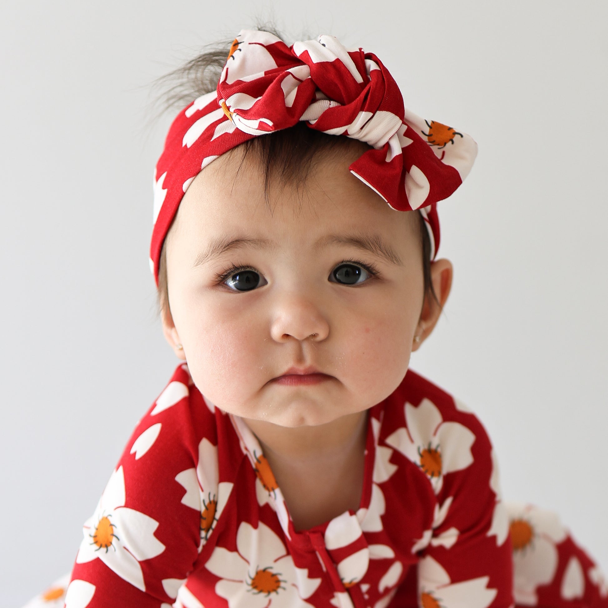 A baby wearing the forever french baby Bamboo Head Wrap | Cherry Blossoms, featuring white and orange floral patterns, pairs it with a matching outfit while looking at the camera with a neutral expression.