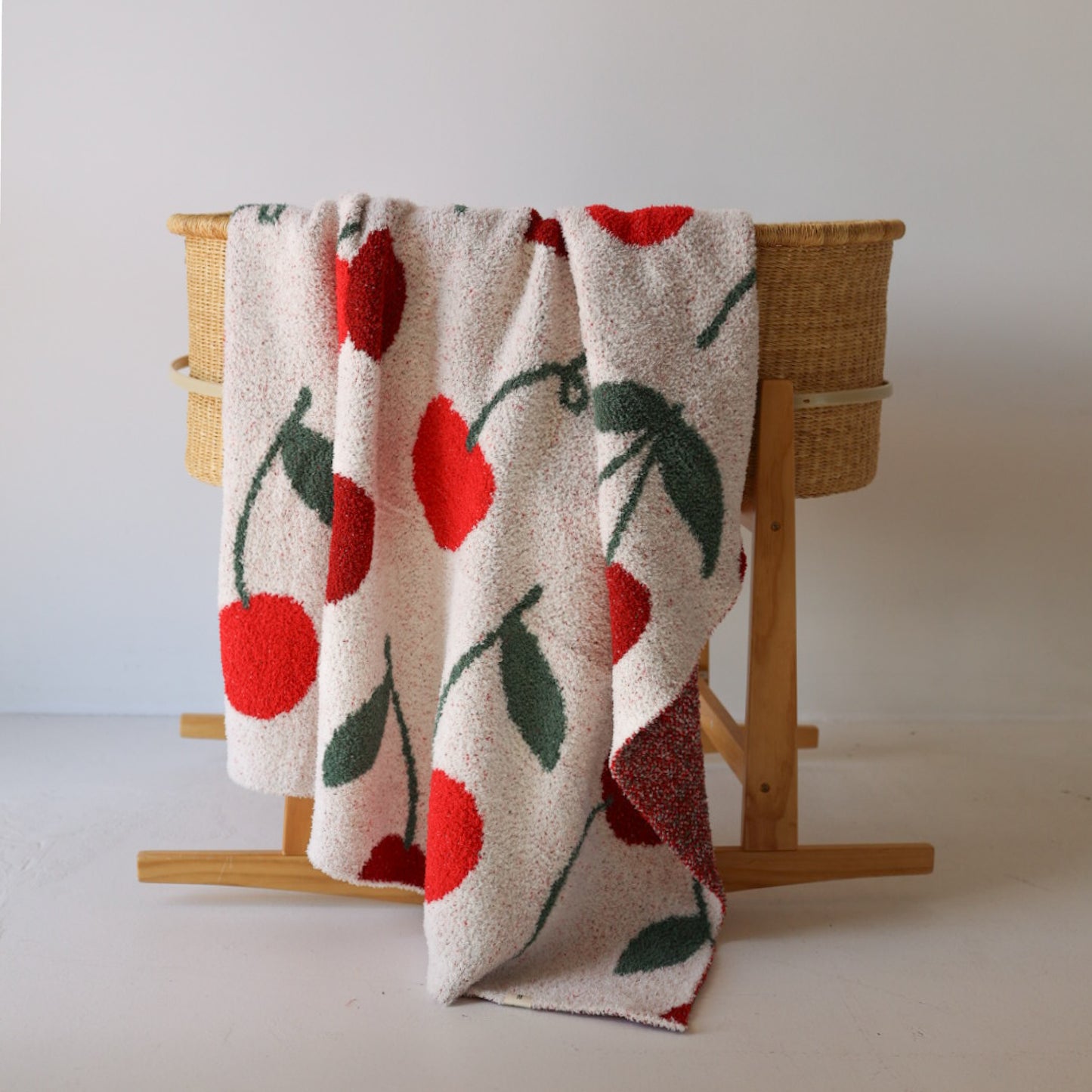 A woven basket stand holds the forever french baby Plush Blanket | Cherry, a white OEKO-TEX certified microfiber throw with large red cherries and green leaves, draped casually over one side against a plain white background.