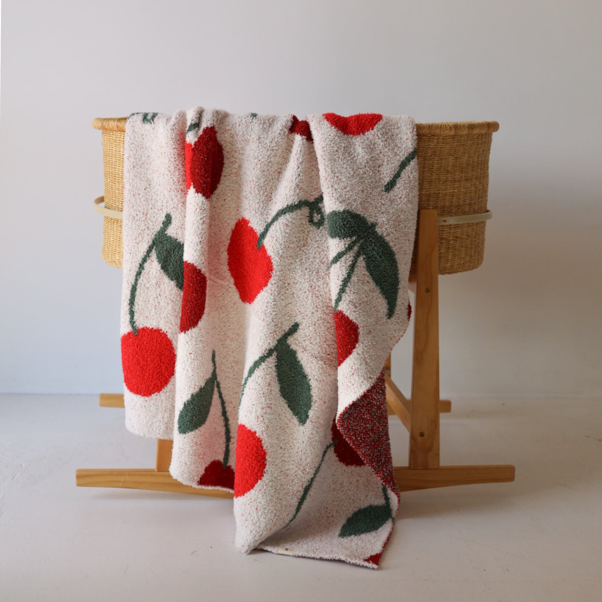 A woven basket stand holds the forever french baby Plush Blanket | Cherry, a white OEKO-TEX certified microfiber throw with large red cherries and green leaves, draped casually over one side against a plain white background.