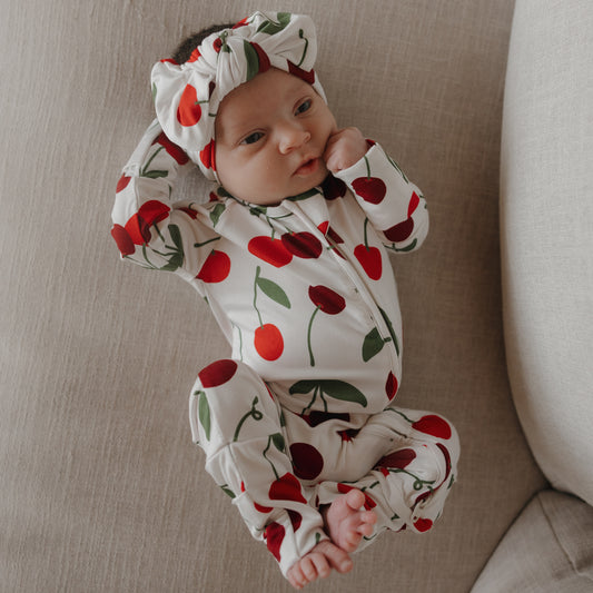 A baby lies on a beige couch, wearing forever french baby's Bamboo Zip Pajamas | Cherry—hypoallergenic white pajamas with a red cherry pattern—and looks up, one hand touching their face and the other arm bent above their head.