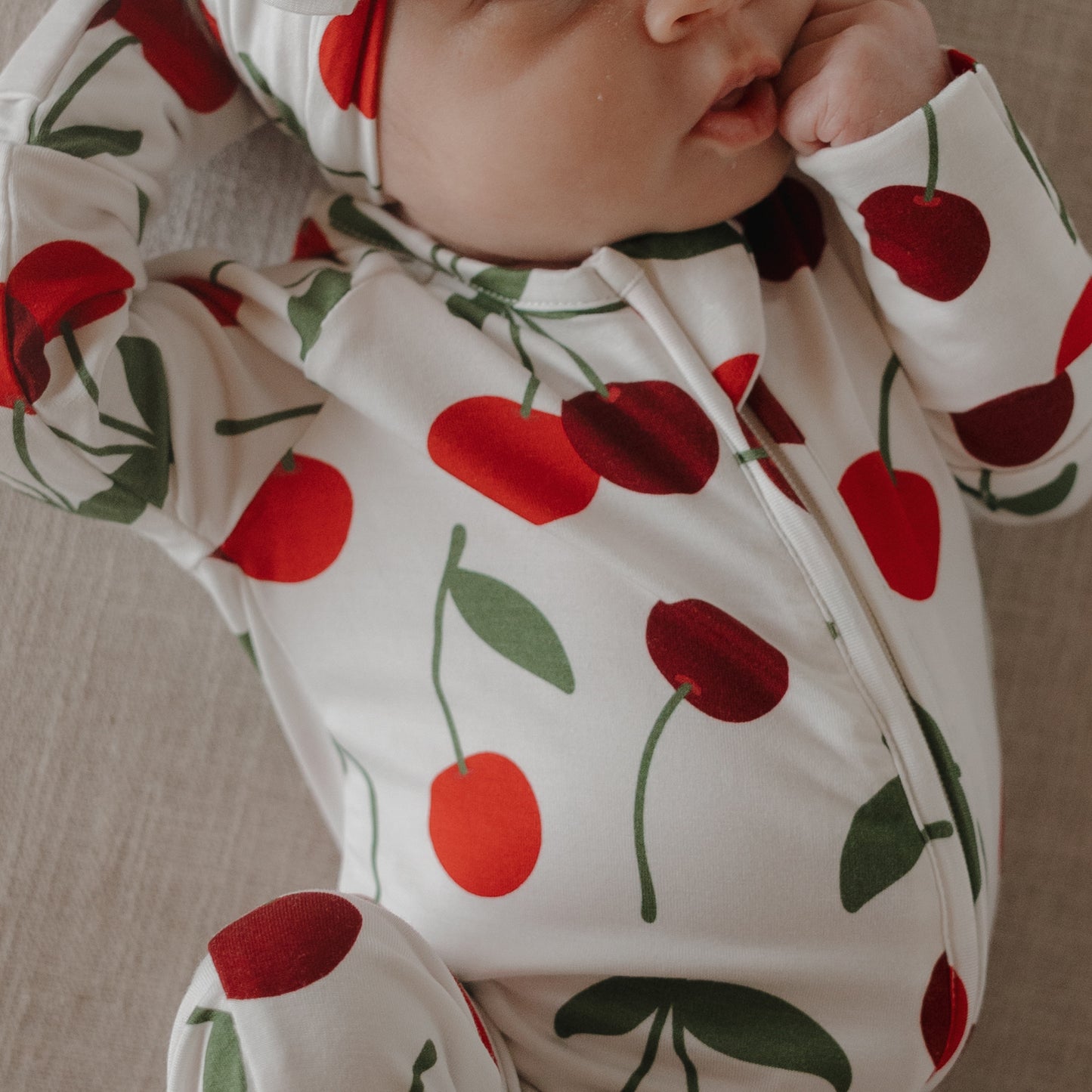 A baby in forever french baby's hypoallergenic Bamboo Zip Pajamas | Cherry—a white onesie and hat with red cherries and green stems—lies on a light surface, one hand touching their face.