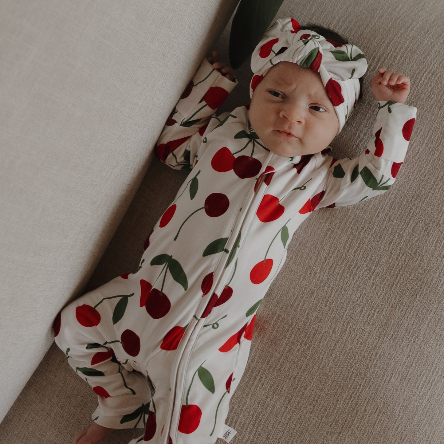 A baby lies on a beige surface, wearing forever french baby's Bamboo Zip Pajamas | Cherry—a white bamboo onesie and matching headband with red cherry and green leaf print—looking upward with one arm raised.