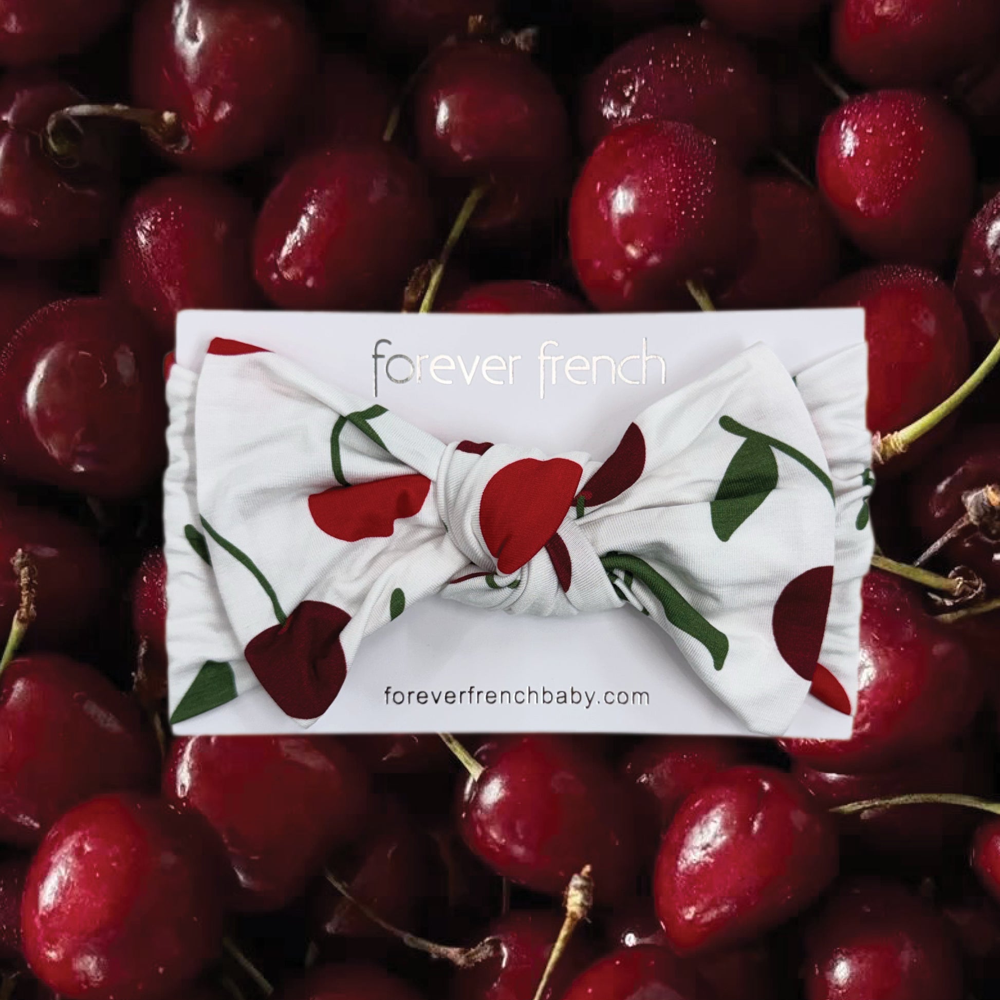 The forever french baby Bamboo Head Wrap | Cherry, featuring a red cherry pattern, is displayed on branded packaging and set on a pile of fresh cherries.