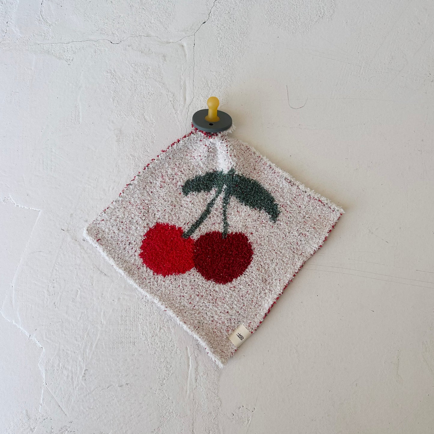 The Lovey | Cherry by forever french baby is a square white security blanket featuring a textured red cherry and green stem design, displayed on a wall from a round hook with a yellow peg.