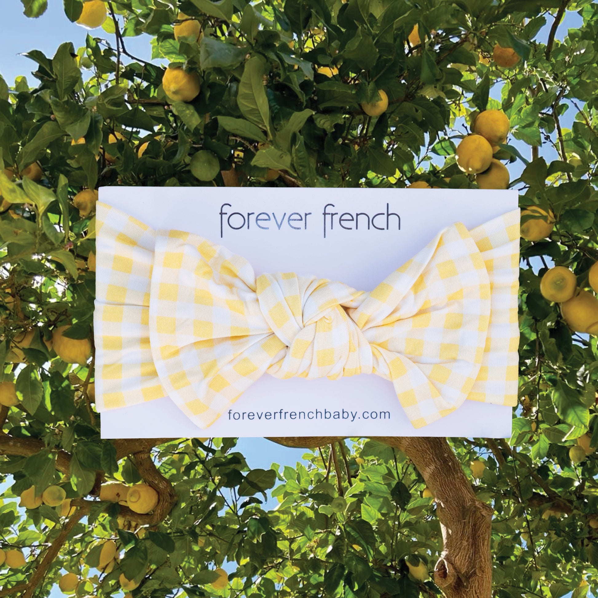 The Bamboo Head Wrap | Citron by forever french baby, a yellow and white gingham baby headband, is shown on a branded card in front of a lemon tree with bright yellow lemons.