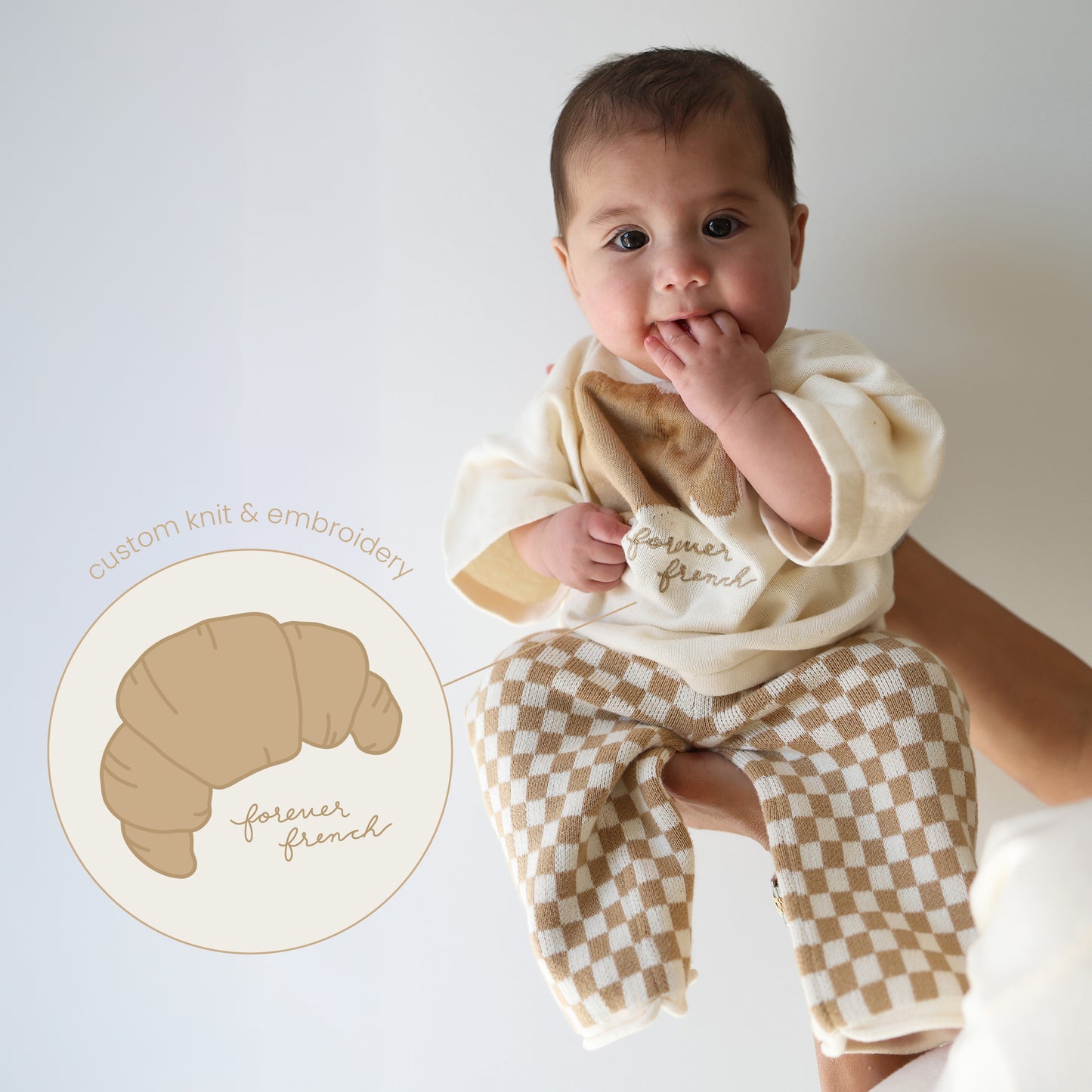 A baby in the forever french baby Knit Pant Set | Croissants—featuring a cream sweater with a croissant design and checked knit pants—is held by an adult. An illustrated croissant with "forever french" and "custom knit & embroidery" appears nearby.