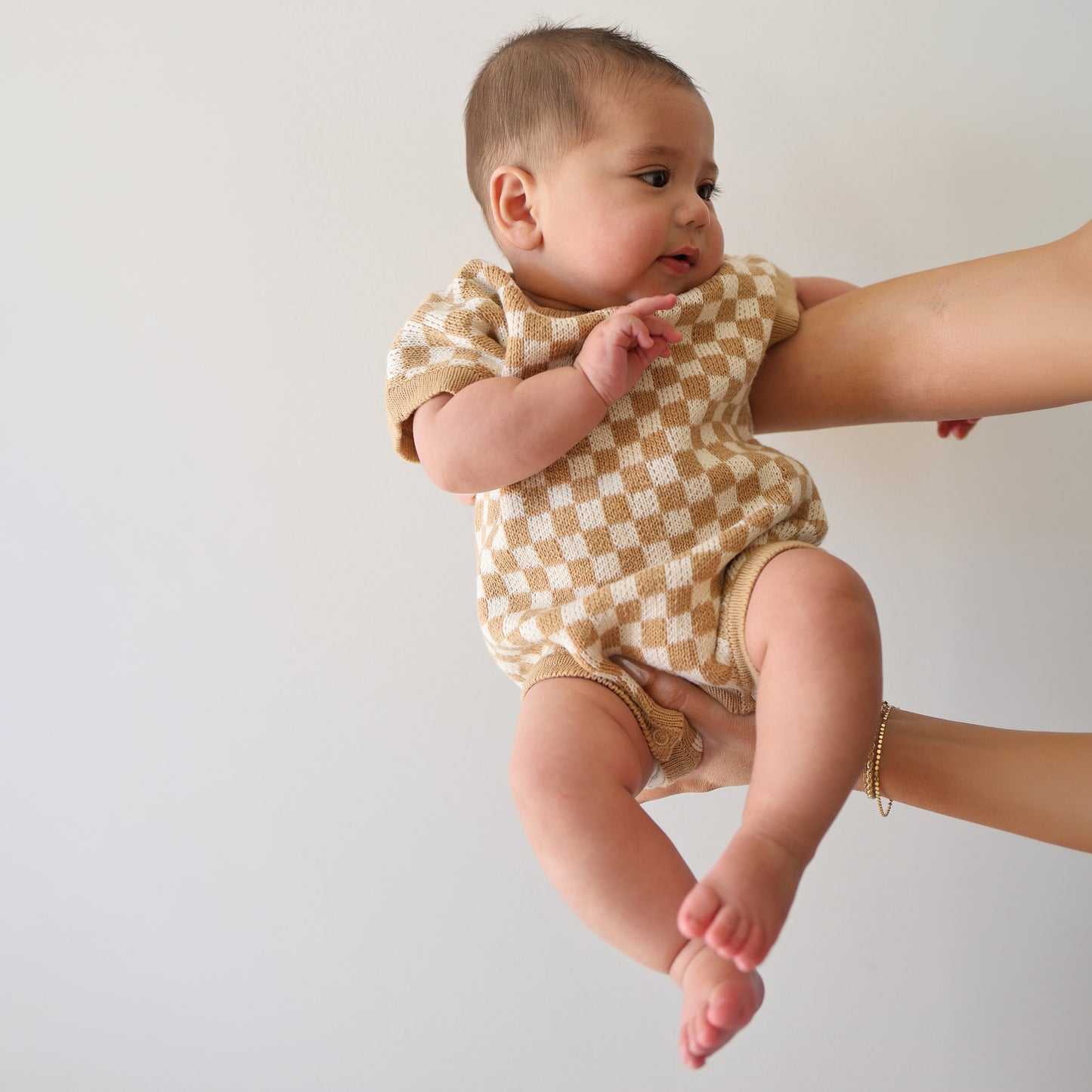 A baby wears the forever french baby Knit Romper | Latte Check, featuring a beige and white checkerboard pattern, while being held by an adult against a plain white background, looking to the side with one arm raised.