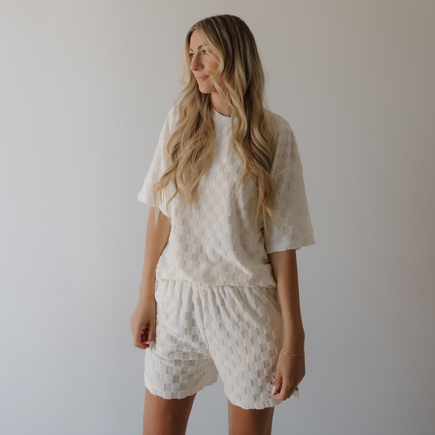 A woman with long, wavy blonde hair wears the Women's Terry Short Set in Coconut Milk by forever french baby. She stands against a plain light background, looking to the side with a relaxed expression.