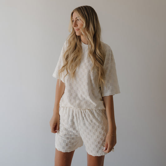 A woman with long, wavy blonde hair wears the Women's Terry Short Set in Coconut Milk by forever french baby. She stands against a plain light background, looking to the side with a relaxed expression.