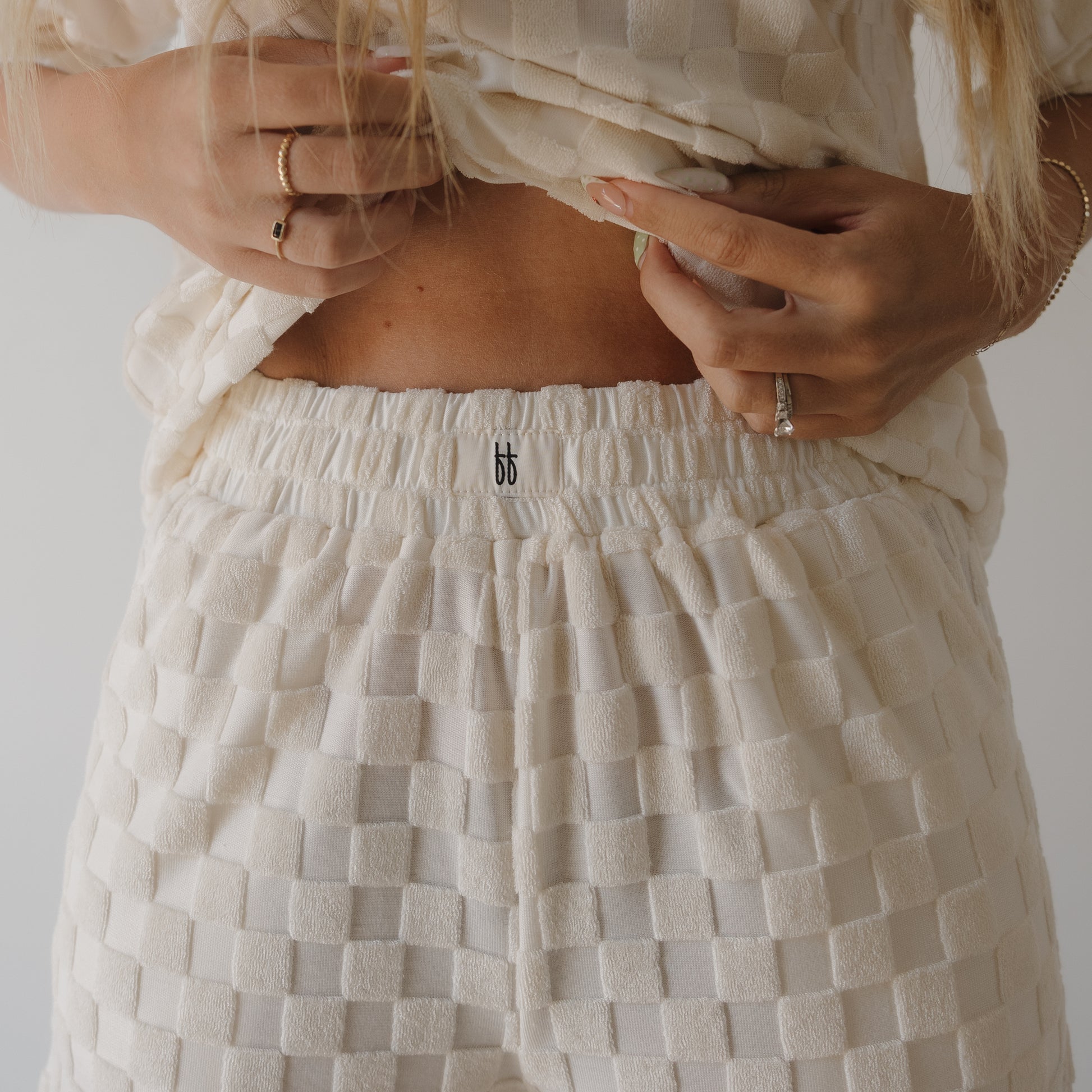 Wearing the Women's Terry Short Set in Coconut Milk by forever french baby, a long-haired person lifts their textured top to reveal the elastic waistband of the matching shorts, styled with rings for an effortlessly cozy look.
