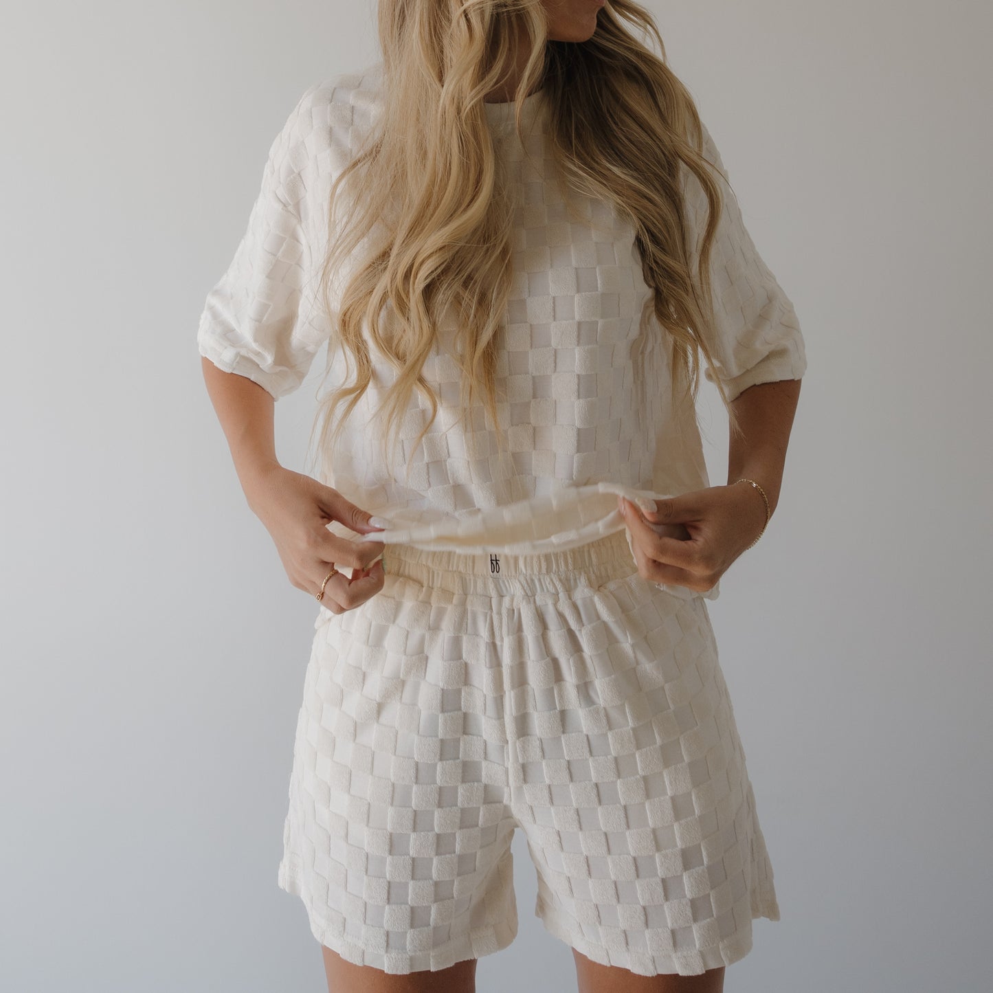 A person with long wavy blonde hair wears the forever french baby Women's Terry Short Set in Coconut Milk, standing against a light background and adjusting the hem—ideal for comfortable loungewear.