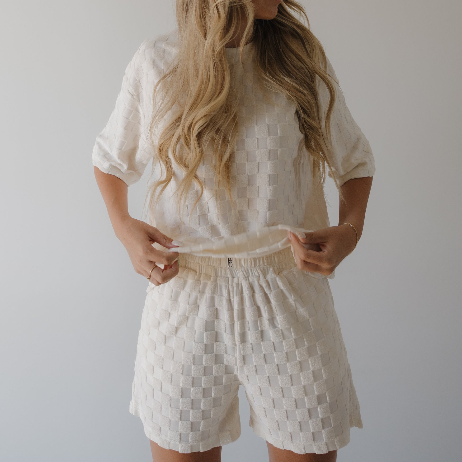 A person with long wavy blonde hair wears the forever french baby Women's Terry Short Set in Coconut Milk, standing against a light background and adjusting the hem—ideal for comfortable loungewear.