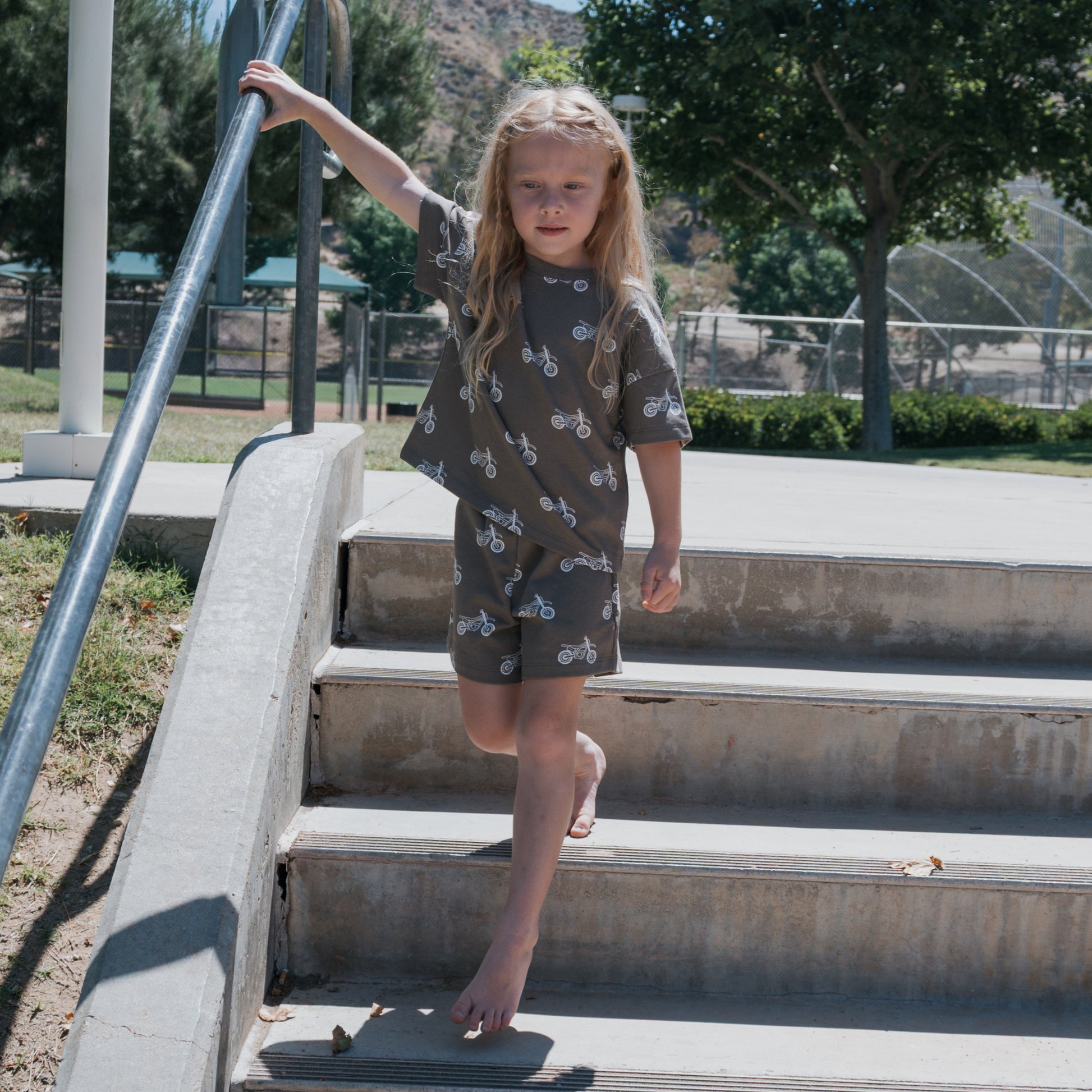 A young child with long blonde hair, barefoot, walks down concrete steps outdoors holding a metal handrail. They wear the forever french baby French Terry Short Set | Dirt Bike. Trees and grass are visible in the background.