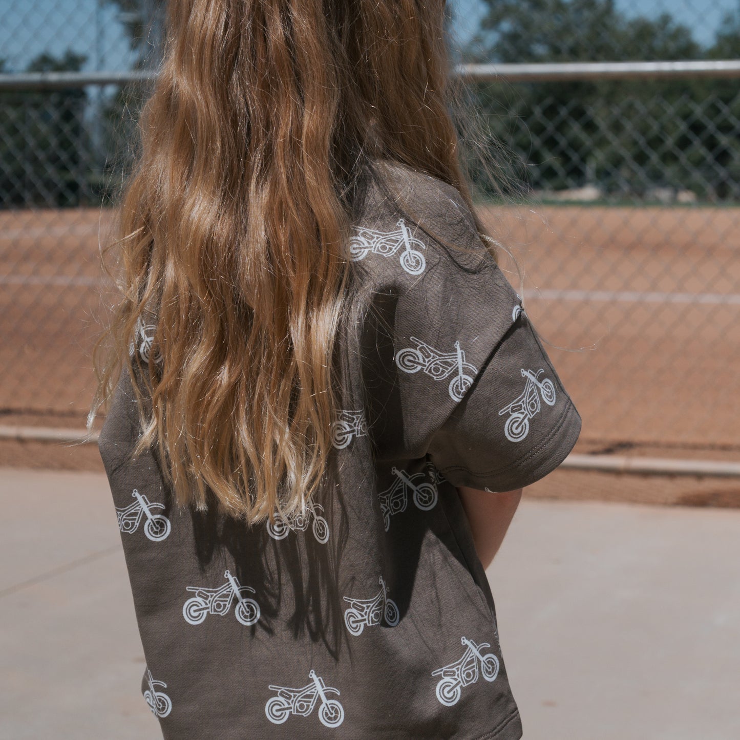 A child with long, wavy blonde hair stands outdoors, facing away in a forever french baby French Terry Short Set | Dirt Bike, featuring white motorcycle prints. A chain-link fence and baseball diamond are visible in the background.