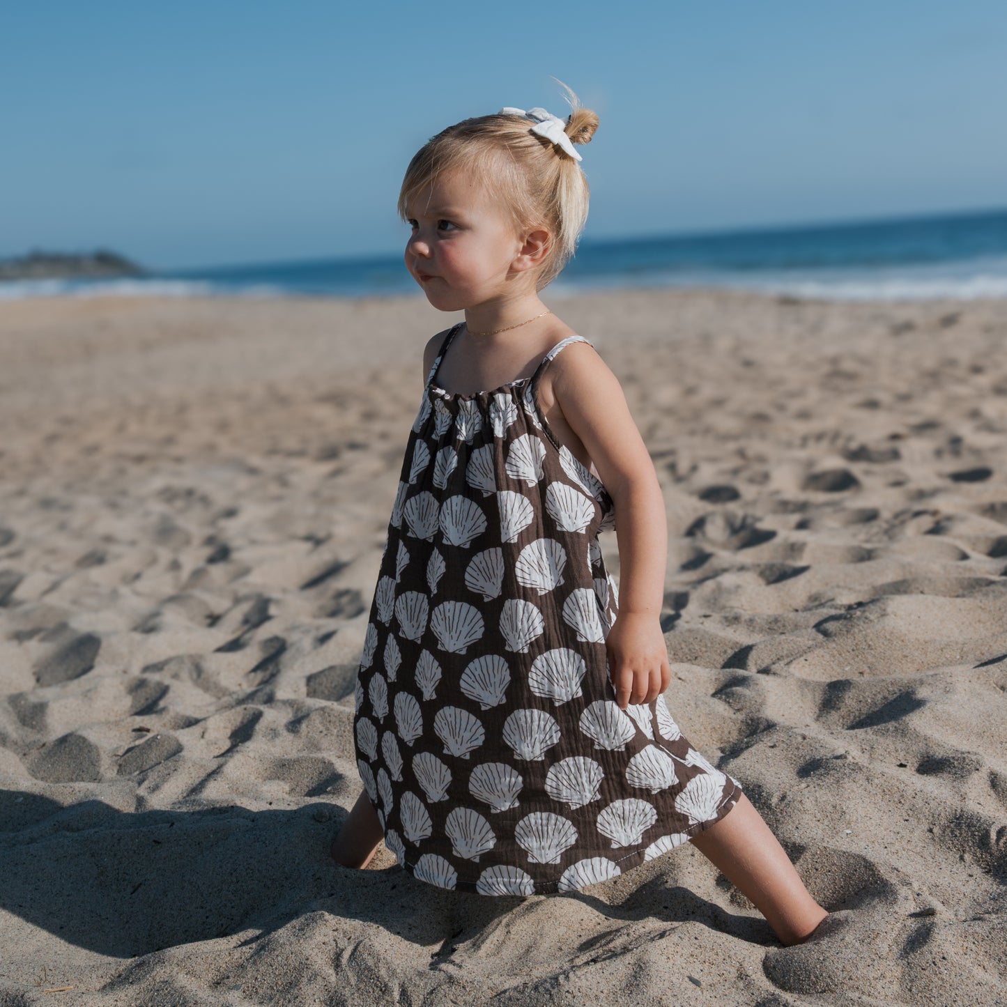A young blond-haired child with a bun stands barefoot on the sand, wearing the forever french baby Child Muslin Sleeveless Dress | Seashells, with the ocean and blue sky in the background.