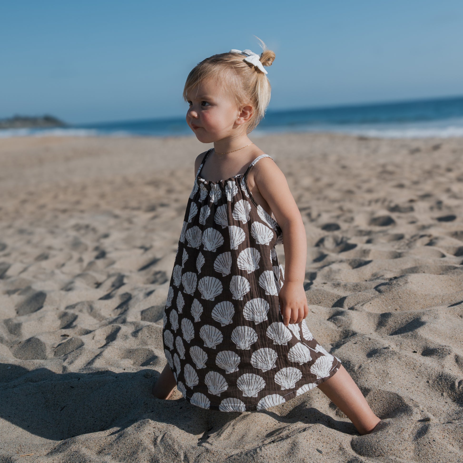 A young blond-haired child with a bun stands barefoot on the sand, wearing the forever french baby Child Muslin Sleeveless Dress | Seashells, with the ocean and blue sky in the background.
