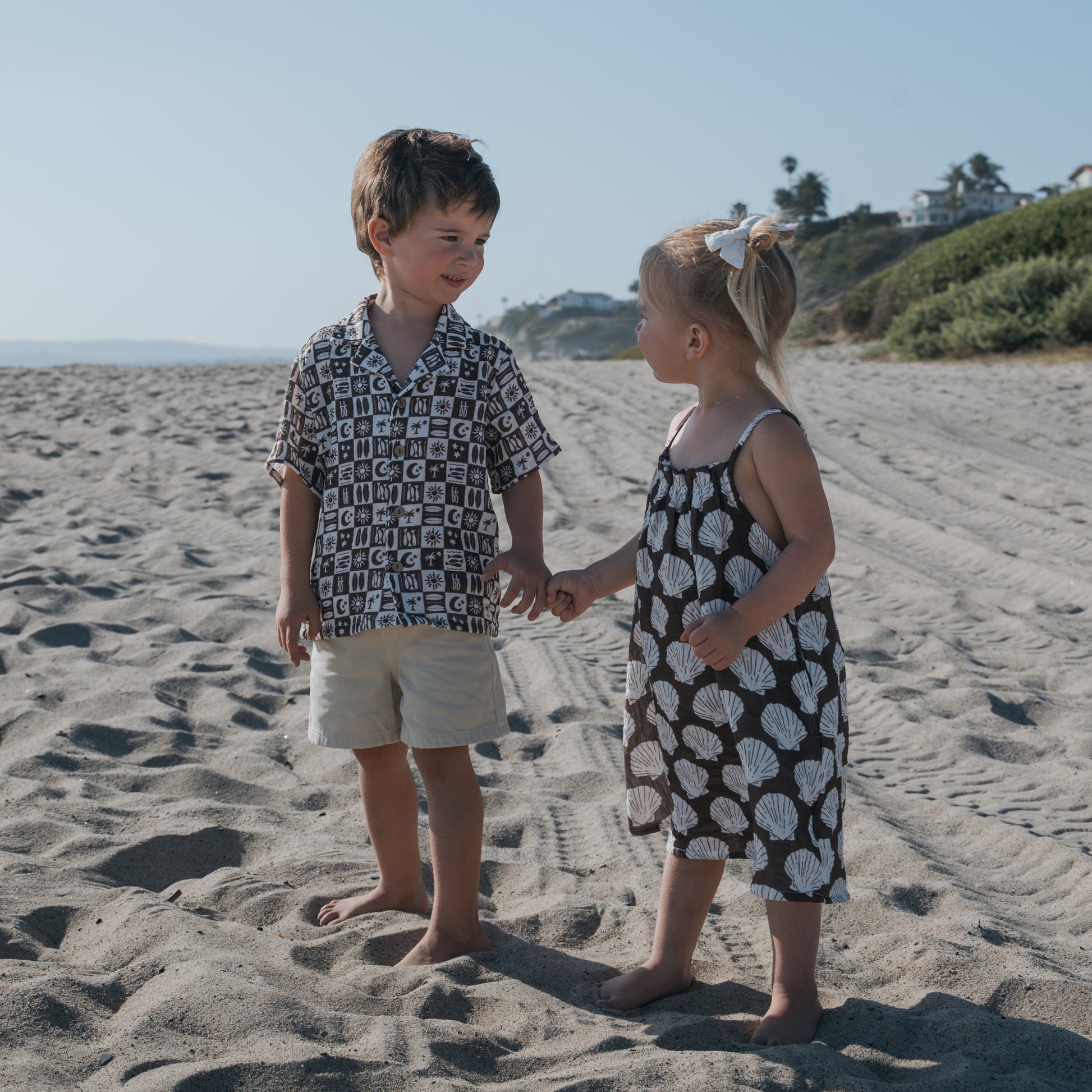A young boy and girl smile at each other, holding hands on a sunny beach. The girl wears the forever french baby Child Muslin Sleeveless Dress | Seashells, with houses and greenery in the background.