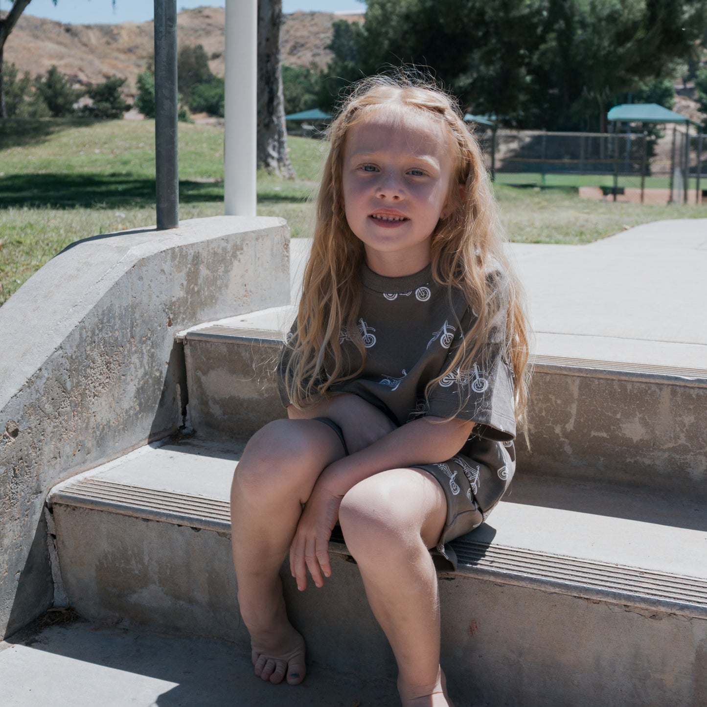 A young child with long blond hair sits on concrete steps in a park, smiling at the camera. They're wearing the forever french baby French Terry Short Set | Dirt Bike. Trees and a playground appear in the background.