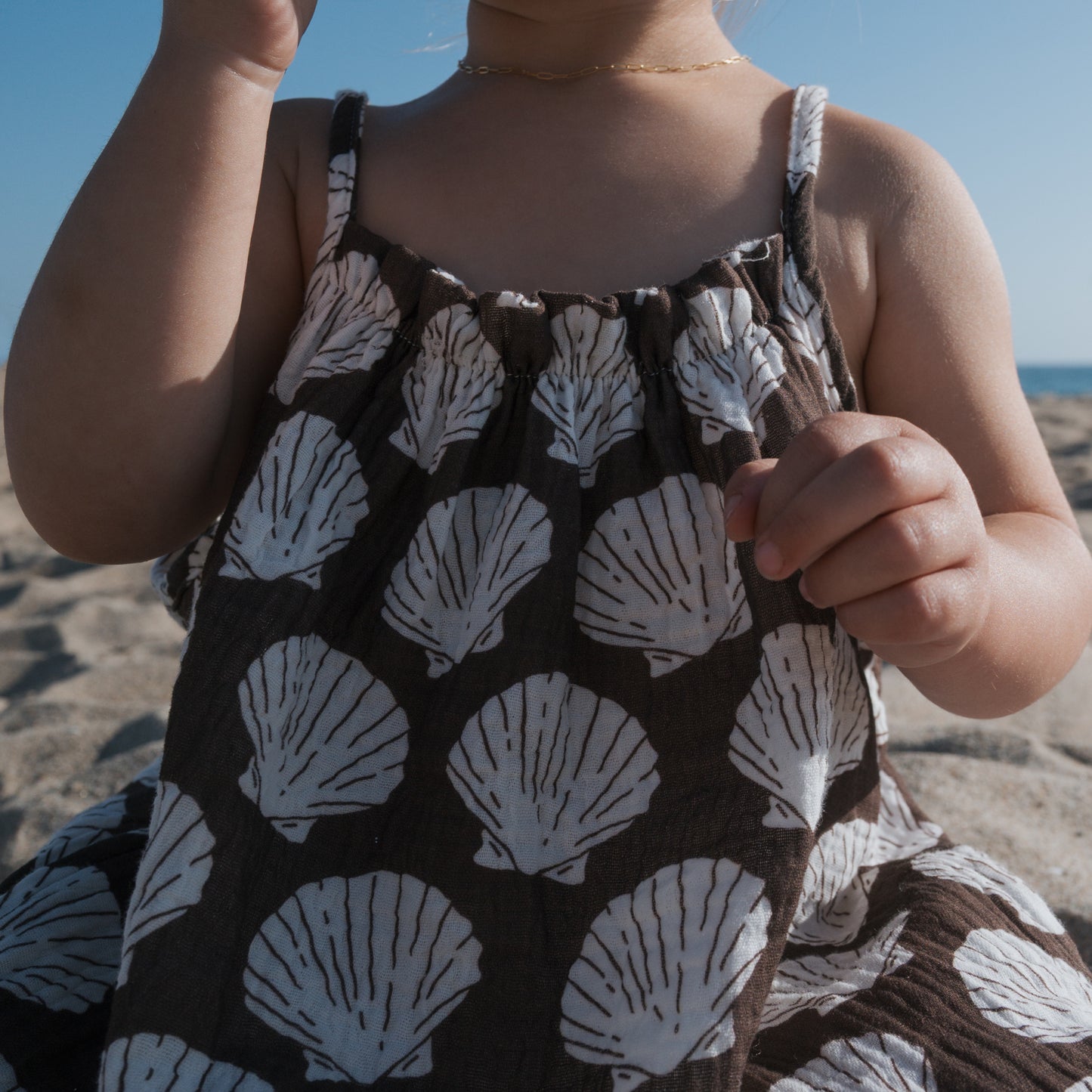 A child in a forever french baby Muslin Sleeveless Dress | Seashells sits on a sandy beach, the ocean behind her—an ideal pick for coordinated family outfits by the shore.